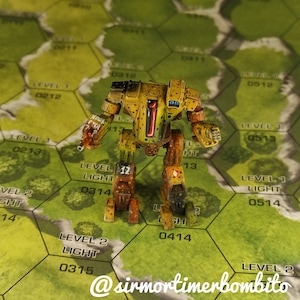 Może przedstawiać: Miniaturowy robot pomalowany na żółto i brązowo z numerem "1" na klatce piersiowej, stojący na zielono-brązowej sześciokątnej planszy do gry z numerami i tekstem.