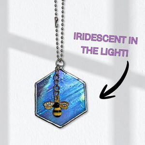 May include: A hexagonal stained glass suncatcher with iridescent blue and purple hues. A small, gold-colored bee charm with rhinestone wings hangs below. The text "IRIDESCENT IN THE LIGHT!" is visible.