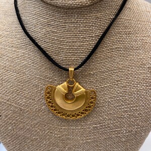 May include: A gold pendant necklace with a large, intricate, circular design. The pendant is suspended from a black cord.