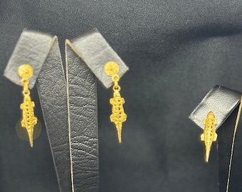 May include: Gold-colored dangle earrings featuring a lizard design. The earrings have a small round stud and a detailed lizard pendant. The earrings are displayed on black earring stands against a dark background.