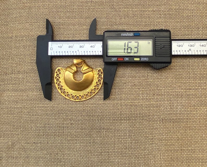 May include: A gold-colored metal pendant in the shape of a crescent moon with a circular cutout in the center. The pendant is 1.63 inches in diameter and has a detailed design with a beaded border.