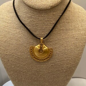 May include: A gold necklace with a large, intricate pendant. The pendant is shaped like a crescent moon with a circular opening in the center. The necklace is on a black cord.