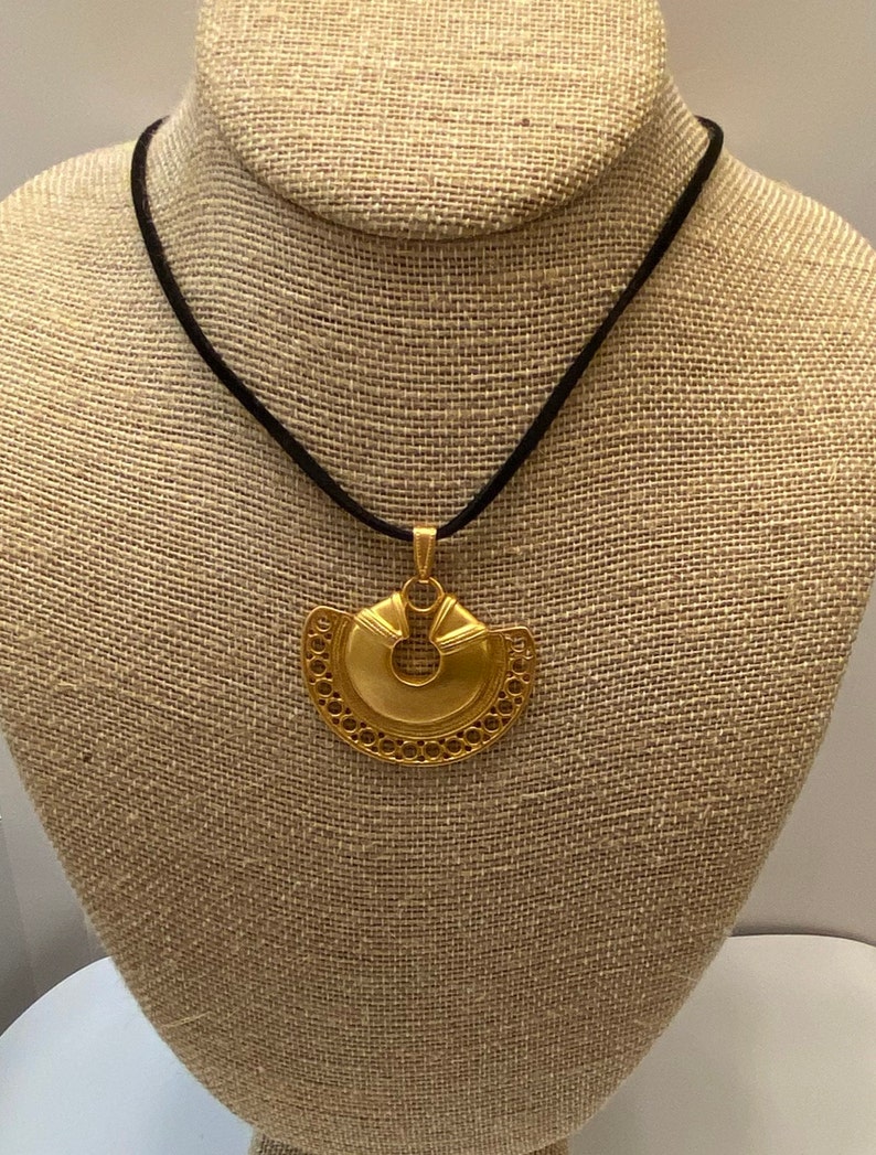 May include: A gold necklace with a large, intricate pendant. The pendant is shaped like a crescent moon with a circular opening in the center. The necklace is on a black cord.