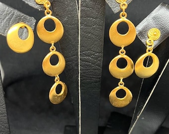 May include: Gold-toned earrings with a circular design. One pair features a stud with a small floral detail, and three graduated open circles hanging below. Another pair has a single open circle stud. The earrings are displayed on black stands.