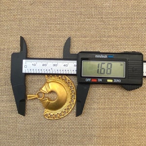 May include: A gold-colored metal pendant with a circular design and a hole in the center. The pendant is being measured with a caliper, which reads 1.68 inches.