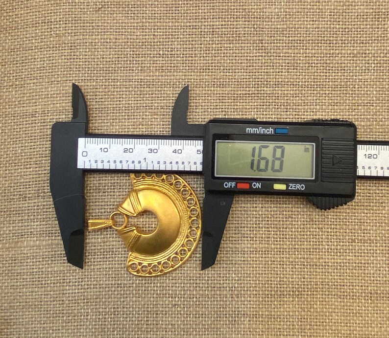 May include: A gold-colored metal pendant with a circular design and a hole in the center. The pendant is being measured with a caliper, which reads 1.68 inches.