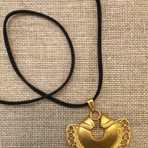 May include: A gold pendant necklace with a large, intricate, circular design. The pendant is attached to a black cord.
