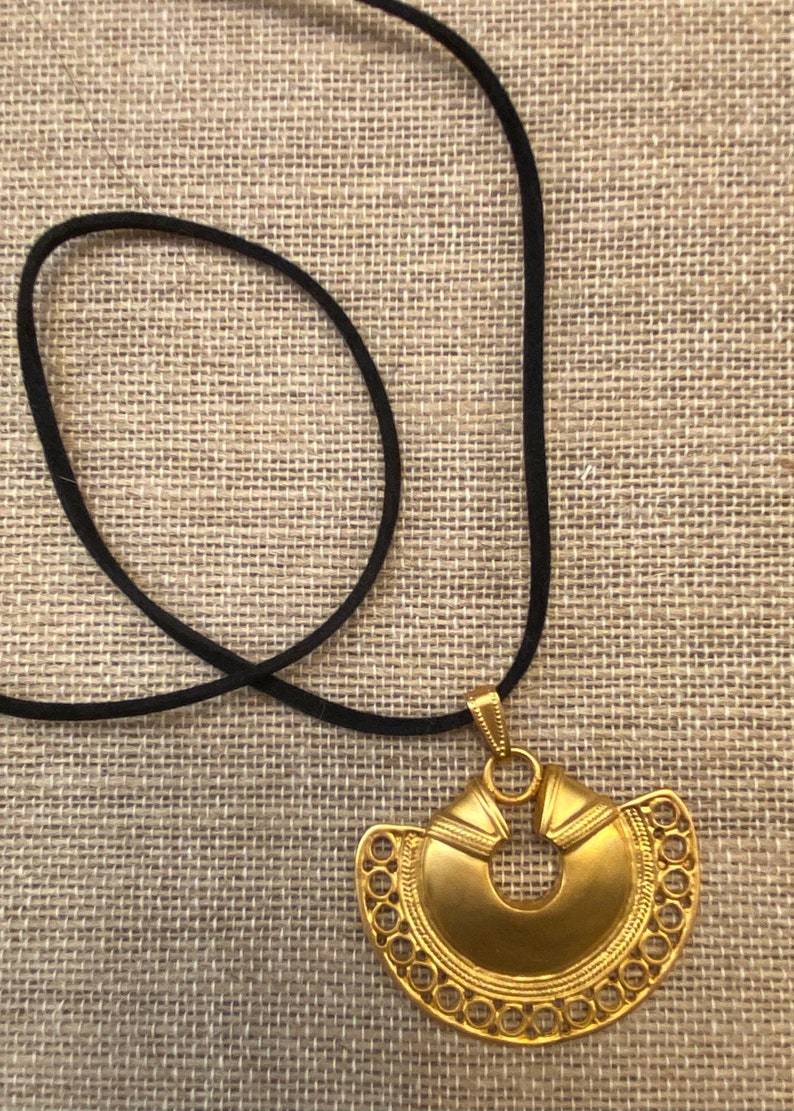 May include: A gold pendant necklace with a large, intricate, circular design. The pendant is attached to a black cord.