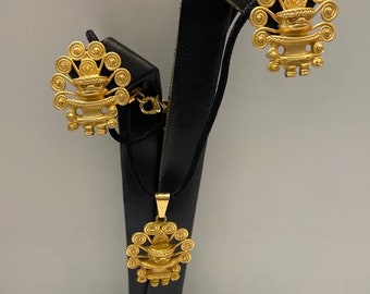 May include: A gold necklace and earrings set featuring a stylized design of a figure with a crown and a circular pattern. The jewelry is displayed on a black stand.