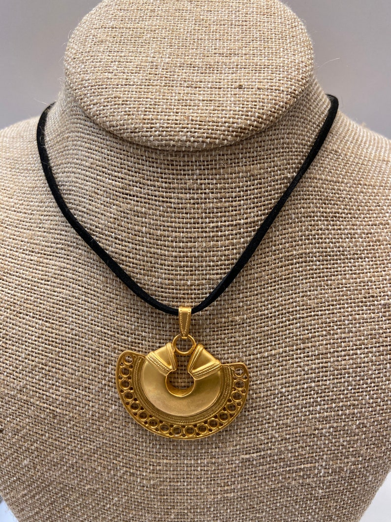 May include: A gold pendant necklace with a large, intricate, circular design. The pendant is suspended from a black cord.