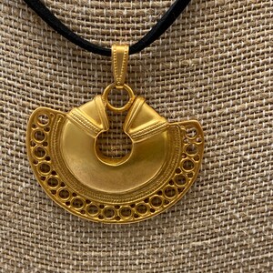 May include: A gold pendant necklace with a large, circular, ornate pendant. The pendant has a circular cutout in the center and is decorated with intricate details. The necklace is on a black cord.