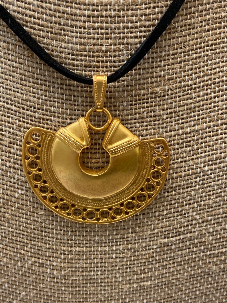 May include: A gold pendant necklace with a large, circular, ornate pendant. The pendant has a circular cutout in the center and is decorated with intricate details. The necklace is on a black cord.
