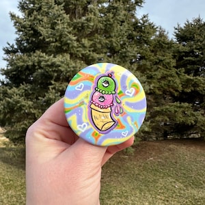 May include: A colorful button with a psychedelic background featuring a swirl pattern and white hearts. The button features a cartoon ice cream cone with three scoops of ice cream, each with an eye. The ice cream is green, pink, and green. The cone is yellow and brown with a cherry on top.