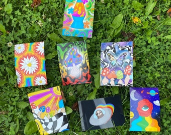 Op de afbeelding: Zeven kleurrijke psychedelische kunstprints met verschillende ontwerpen, waaronder een lava lamp, paddenstoelen, een vlinder, een zon, een luiaard in een ruimteschip en een roos met een oog. De prints zijn op een groene grasachtige achtergrond.