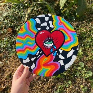May include: A round canvas painting with a red heart with a green eye and blue tears. The heart is surrounded by rainbow-colored swirls and a black and white checkered pattern. The painting is signed "glak.14".