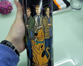 Puede incluir: Un vaso de acero inoxidable negro con un gráfico de cuatro personajes del programa de televisión Supernatural y Scooby Doo. Los personajes están de pie frente a un fondo negro con un diseño de remolino morado. El vaso tiene una pajita de plástico transparente.