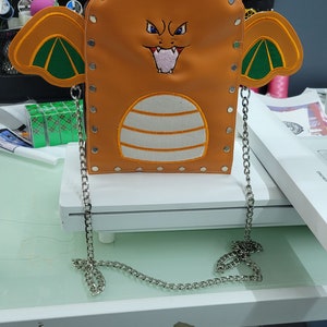 May include: Orange crossbody bag featuring an embroidered cartoon dragon face with a fierce expression. The bag has wings and a silver chain strap. The design includes white and orange stripes and is secured with silver rivets.