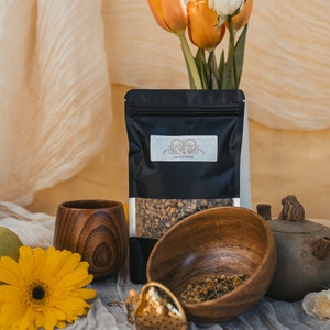 May include: A black pouch of dried herbs labelled "Sacred Herbs" with a wooden bowl filled with loose leaf tea, a wooden cup, a gold tea infuser, and a small ceramic teapot.