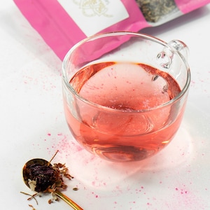 May include: A glass cup filled with pink herbal tea, a pink pouch of loose leaf tea, and a gold spoon with dried tea leaves on a white surface.