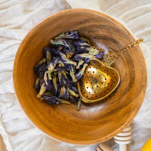 May include: A wooden bowl filled with dried blue butterfly pea flowers and a gold heart-shaped tea infuser with a chain.