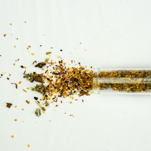Può includere: Un tubetto di vetro trasparente pieno di erbe e spezie essiccate, che si rovesciano su una superficie bianca. Il mix include elementi verdi, marroni e gialli, suggerendo un uso culinario o medicinale. Vista dall'alto.