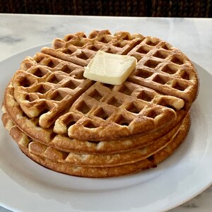 May include: A stack of three golden brown waffles with a pat of butter on top. The waffles are on a white plate.