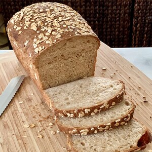 May include: A loaf of freshly baked bread, partially sliced, sits on a wooden cutting board. The bread has a golden-brown crust and is topped with oats. Three slices are visible, showcasing the soft interior. A serrated knife rests beside the loaf.