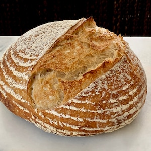 May include: A freshly baked loaf of sourdough bread with a golden brown crust and a slightly cracked top. The bread has a rustic appearance and is dusted with flour.