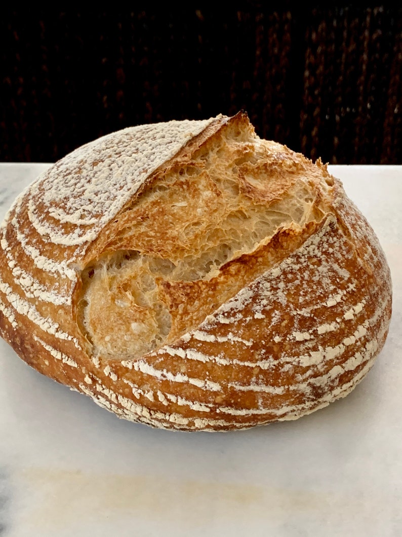 May include: A freshly baked loaf of sourdough bread with a golden brown crust and a slightly cracked top. The bread has a rustic appearance and is dusted with flour.