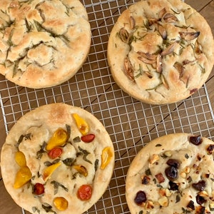 May include: Four different types of focaccia bread on a wire rack. The focaccia on the top left is topped with rosemary, the focaccia on the top right is topped with garlic, the focaccia on the bottom left is topped with cherry tomatoes and basil, and the focaccia on the bottom right is topped with olives, feta cheese, and herbs.