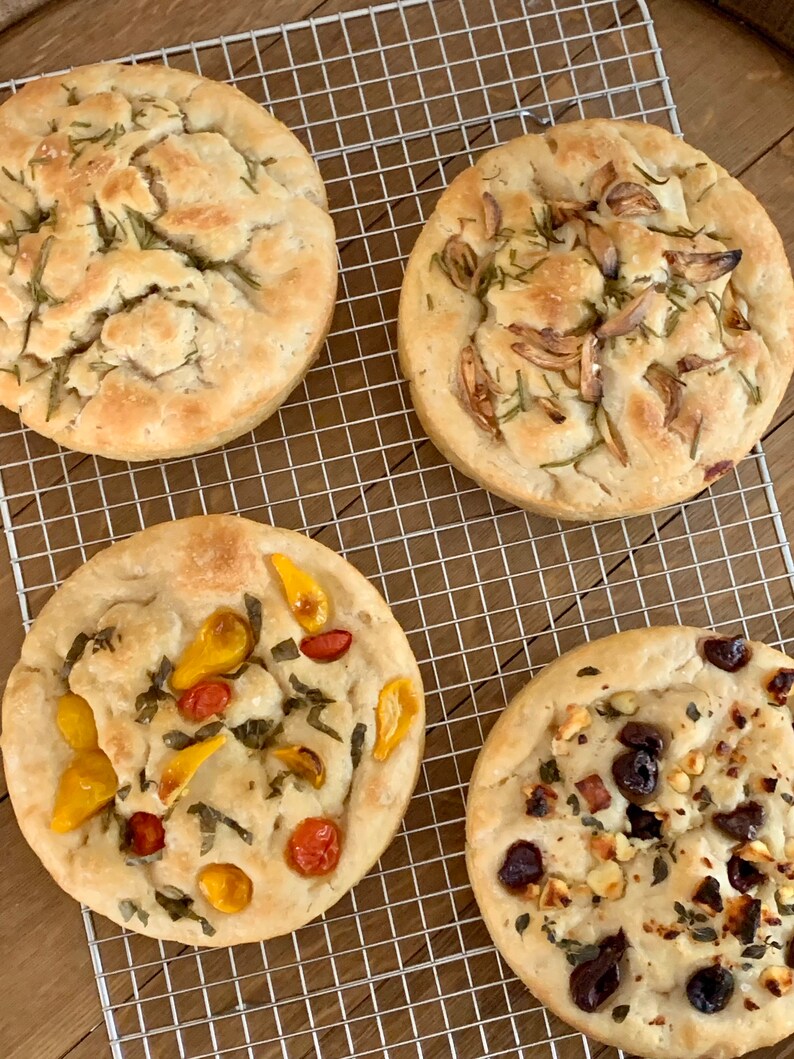 May include: Four different types of focaccia bread on a wire rack. The focaccia on the top left is topped with rosemary, the focaccia on the top right is topped with garlic, the focaccia on the bottom left is topped with cherry tomatoes and basil, and the focaccia on the bottom right is topped with olives, feta cheese, and herbs.