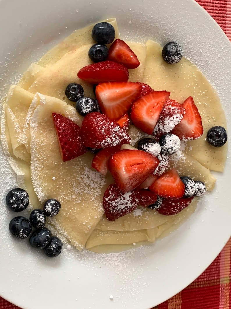 May include: A plate of thin, white crepes topped with fresh strawberries and blueberries. The crepes are dusted with powdered sugar.