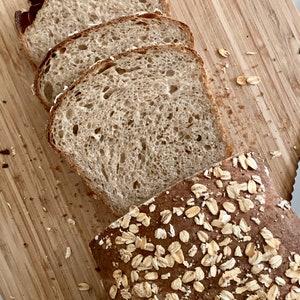 May include: A loaf of bread sliced open, revealing a light brown crumb with a slightly chewy texture. The bread is topped with rolled oats.