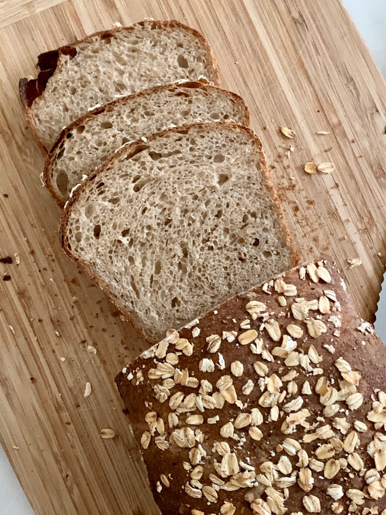 May include: A loaf of bread sliced open, revealing a light brown crumb with a slightly chewy texture. The bread is topped with rolled oats.