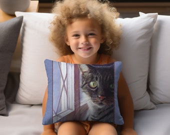 May include: A child sits on a couch holding a throw pillow with a cat looking out a window. The cat is gray and white with green eyes. The pillow is blue with a white window and a gray and white cat.