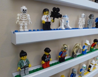 Può includere: Scaffali bianchi con una varietà di minifigure Lego esposte su di essi. Le minifigure includono uno scheletro, un uomo con una macchina fotografica, un robot, un uomo in abito, una donna con una camicia verde e un uomo con una giacca nera.