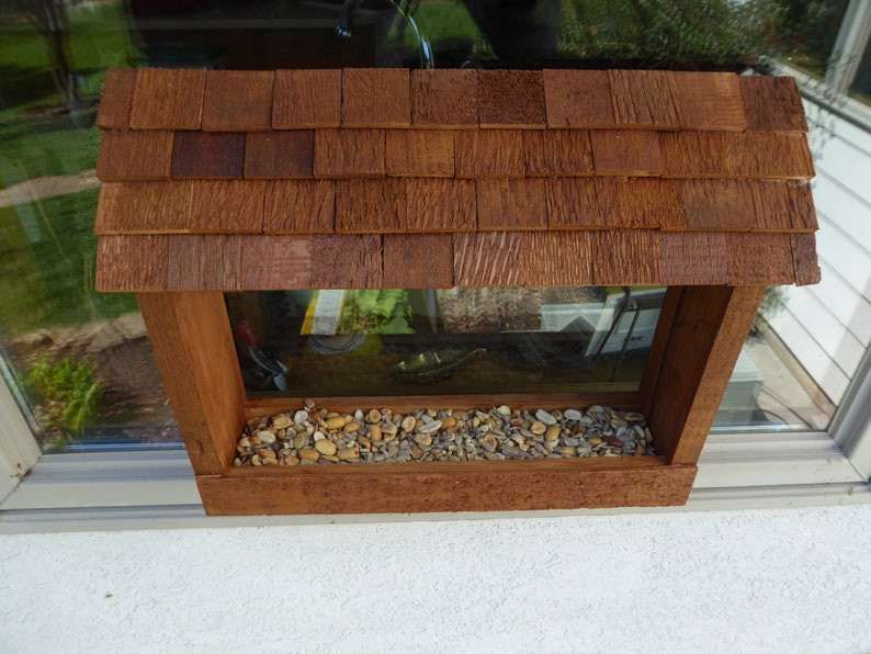 K&ouml;nnte beinhalten: Vogelhaus aus Holz mit Schindeldach und einem transparenten Fenster. Das Futterhaus ist mit kleinen Kieselsteinen gef&uuml;llt und an einer Fensterbank montiert. Das Holz hat eine nat&uuml;rliche, rotbraune Beize.