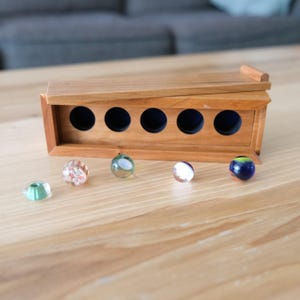 May include: A rectangular wooden box with a slanted lid and five circular openings lined with dark blue fabric. Five glass marbles of varying colors and patterns, including clear, green, white, and blue, are arranged in a row in front of the box.