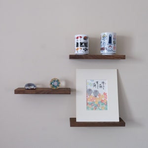 May include: Two wooden shelves with a white ceramic mug with a sushi design, a white ceramic mug with a black and white design, a glass paperweight with a flower design, and a framed print with Japanese calligraphy and a watercolor painting of flowers.