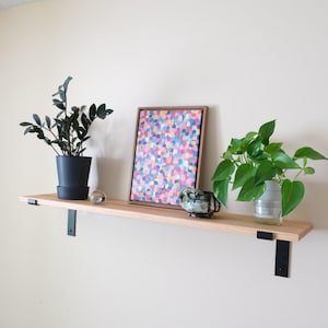 May include: A wooden shelf with black metal brackets, displaying a framed abstract painting, two potted plants, a decorative ceramic bowl, and a clear glass sphere. The painting features colorful brushstrokes.