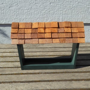 K&ouml;nnte beinhalten: Ein h&ouml;lzernes Vogelfutterhaus mit einem Schindeldach. Das Dach besteht aus kleinen, quadratischen, rotbraunen Holzschindeln. Der Rahmen des Futterhauses ist dunkelgr&uuml;n, mit einer rechteckigen Basis und abgewinkelten St&uuml;tzen.