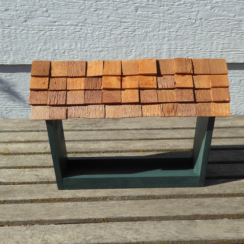 K&ouml;nnte beinhalten: Ein h&ouml;lzernes Vogelfutterhaus mit einem Schindeldach. Das Dach besteht aus kleinen, quadratischen, rotbraunen Holzschindeln. Der Rahmen des Futterhauses ist dunkelgr&uuml;n, mit einer rechteckigen Basis und abgewinkelten St&uuml;tzen.
