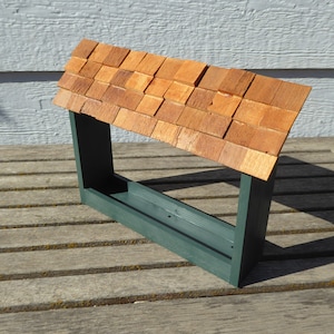 May include: A wooden bird feeder with a green frame and a sloped roof made of small, rectangular cedar shingles. The feeder is designed to be mounted and has a trough for food.