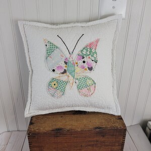Op de afbeelding: Een vierkant wit kussen met een patchwork vlinderontwerp. De vlinder heeft vleugels gemaakt van verschillende stoffen met patronen in roze, groene en blauwe tinten. Het kussen ligt op een houten kist.