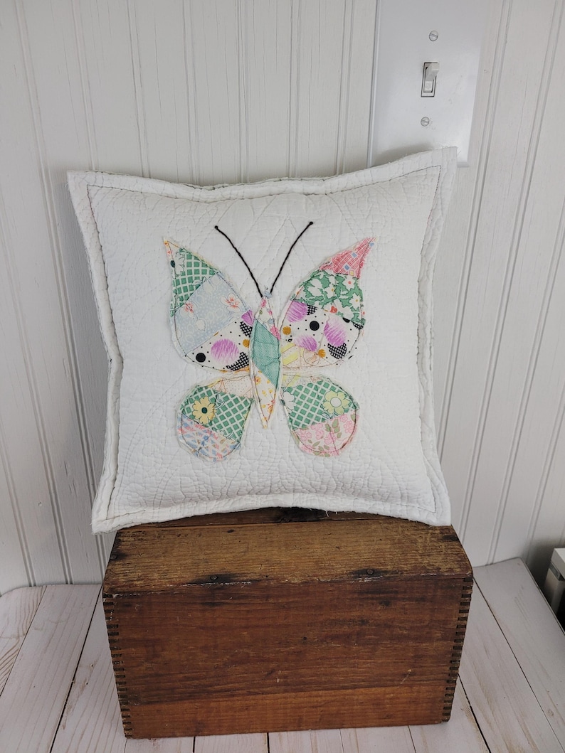 Op de afbeelding: Een vierkant wit kussen met een patchwork vlinderontwerp. De vlinder heeft vleugels gemaakt van verschillende stoffen met patronen in roze, groene en blauwe tinten. Het kussen ligt op een houten kist.