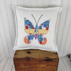 Op de afbeelding: Een vierkant wit kussen met een gewatteerde textuur heeft een vlinderapplicatie. De vlinder heeft blauwe, oranje en geel gekleurde vleugels en een bruine antenne. Het kussen ligt op een houten kist.