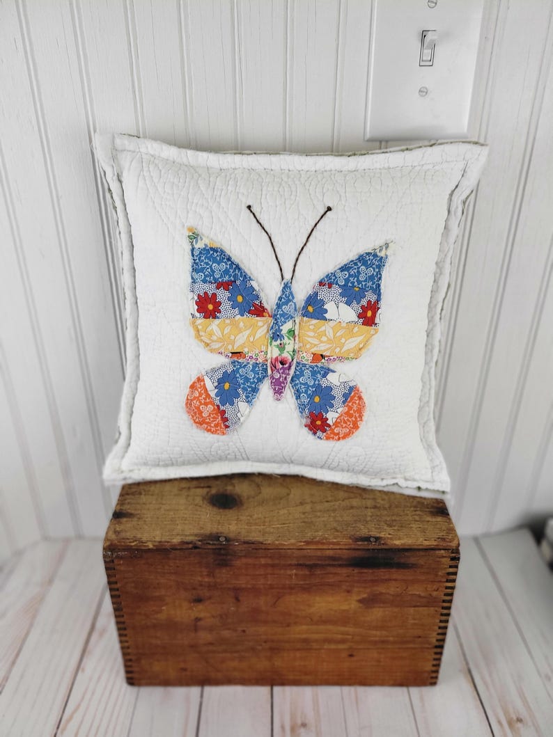 Op de afbeelding: Een vierkant wit kussen met een gewatteerde textuur heeft een vlinderapplicatie. De vlinder heeft blauwe, oranje en geel gekleurde vleugels en een bruine antenne. Het kussen ligt op een houten kist.