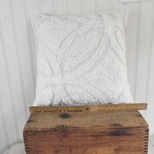 May include: A square, white decorative pillow with a textured, raised leaf pattern. The pillow is placed on a wooden crate. A wooden ruler with the text "The DAKOTA FARMER" is on the crate.