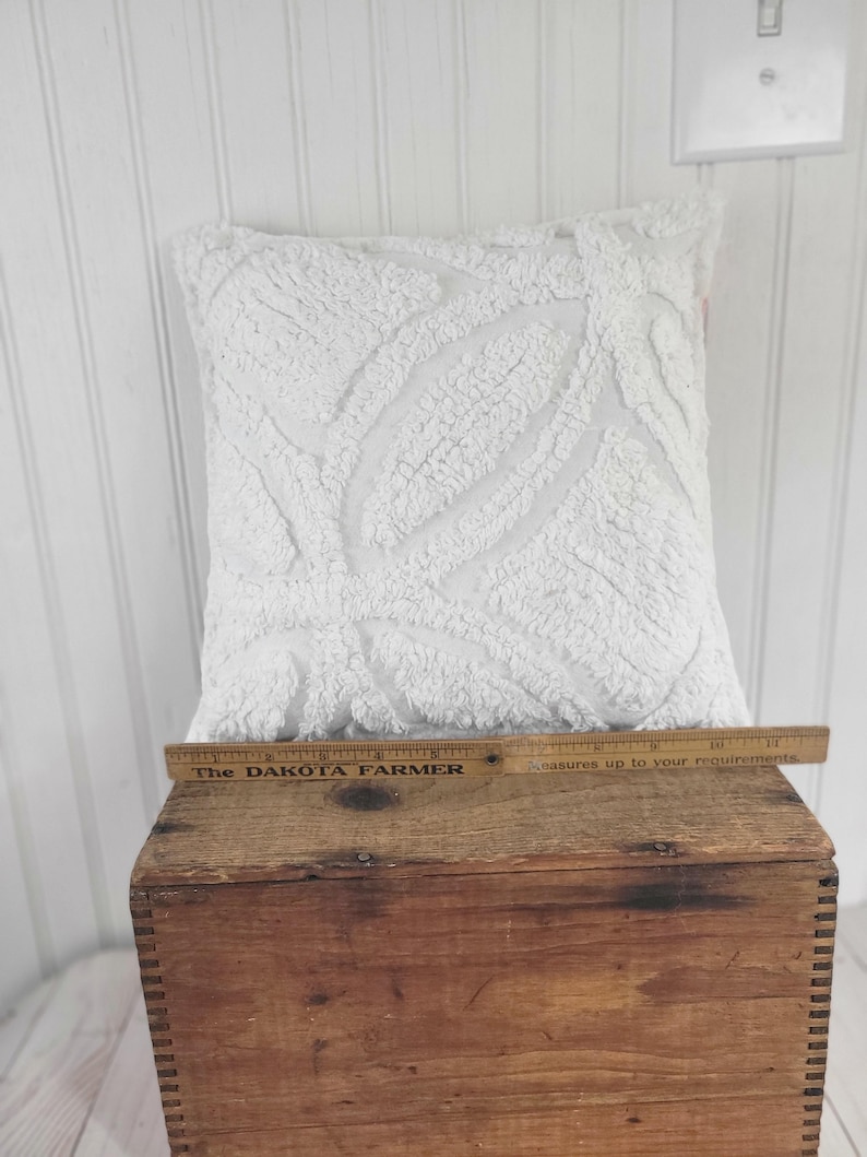 May include: A square, white decorative pillow with a textured, raised leaf pattern. The pillow is placed on a wooden crate. A wooden ruler with the text "The DAKOTA FARMER" is on the crate.