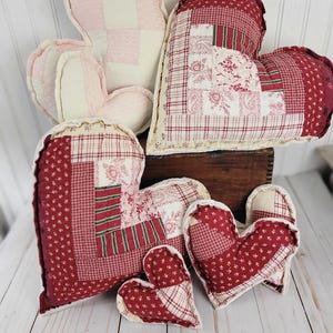 May include: A collection of heart-shaped pillows in various sizes and patterns. The pillows feature patchwork designs in shades of red, pink, and cream. Some have a quilted appearance, while others have a ruffled edge. The pillows are arranged on a wooden surface.